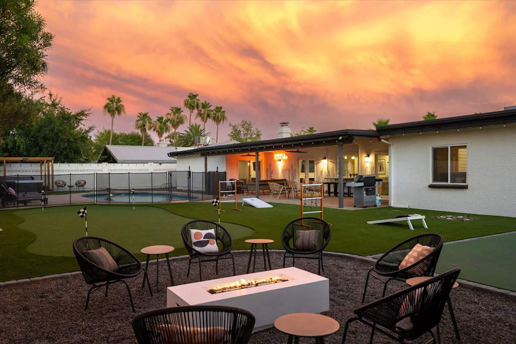 The Sunkiss — backyard fire pit lounge with black chairs, putting green, and pool under a colorful sunset sky