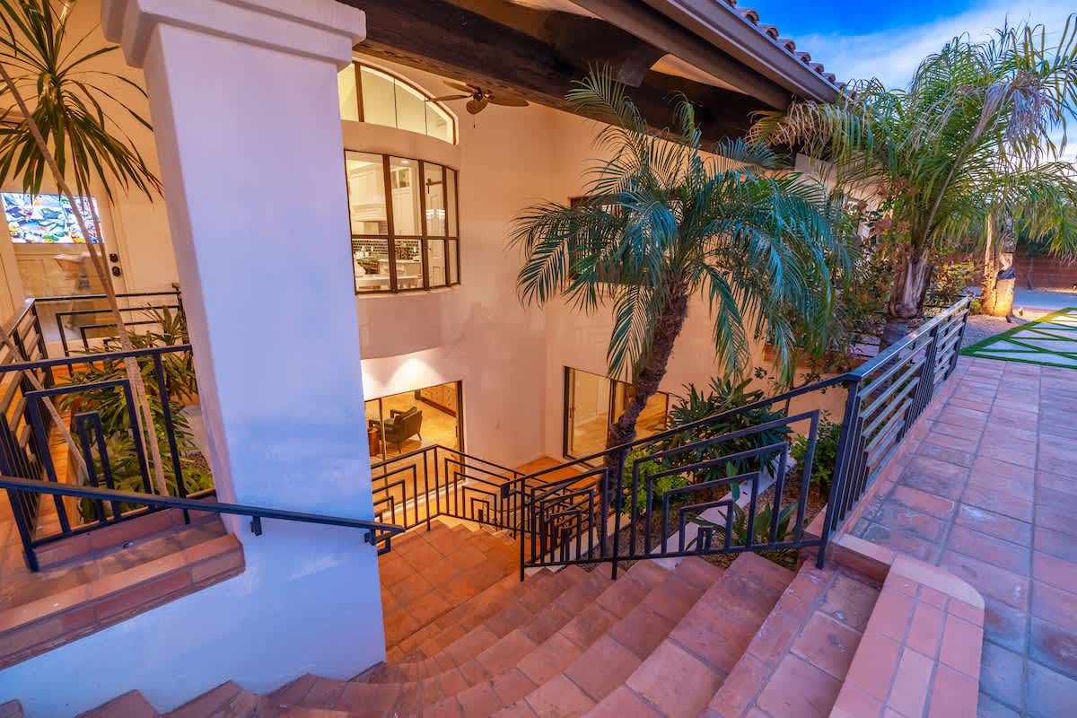 A stunning two-story villa entrance showcasing elegant Spanish-style architecture with terracotta tile steps, lush palm trees, and an inviting courtyard bathed in warm evening light under clear blue skies.