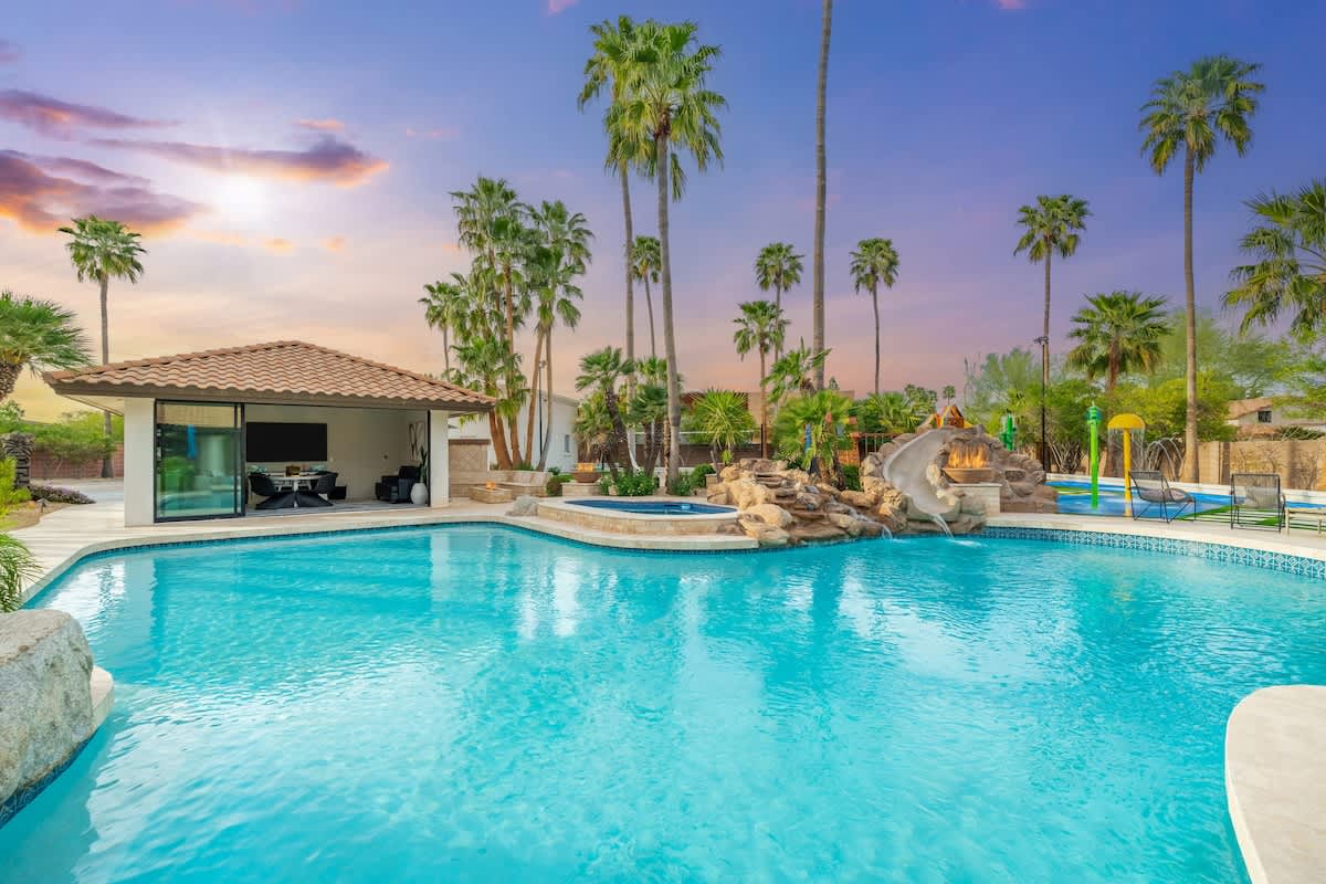 An expansive tropical oasis showcasing a sparkling pool with waterslide, swaying palms, and a shaded cabana area beneath a stunning painted sunset sky.