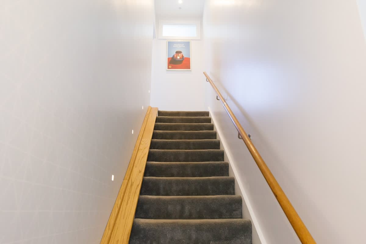 Cedar House: Bright staircase with soft carpeting, wooden handrails, and ambient lighting. A modern and inviting design leads you to the upper level, accented with unique artwork for a welcoming touch.