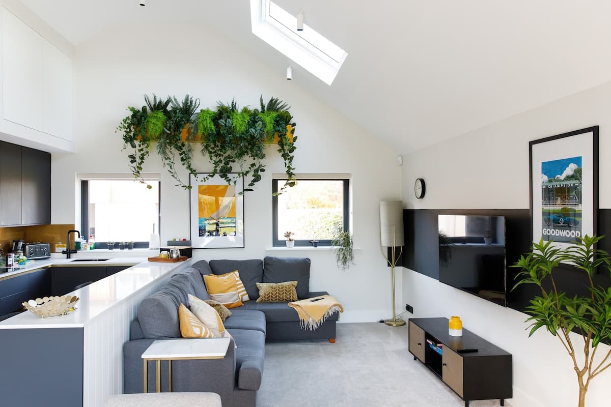 Stylish and cozy living space with a modern L-shaped sofa, smart TV, and vibrant greenery accents. Skylight and large windows fill the room with natural light, creating a bright and inviting atmosphere. Perfect for relaxing or enjoying quality time
