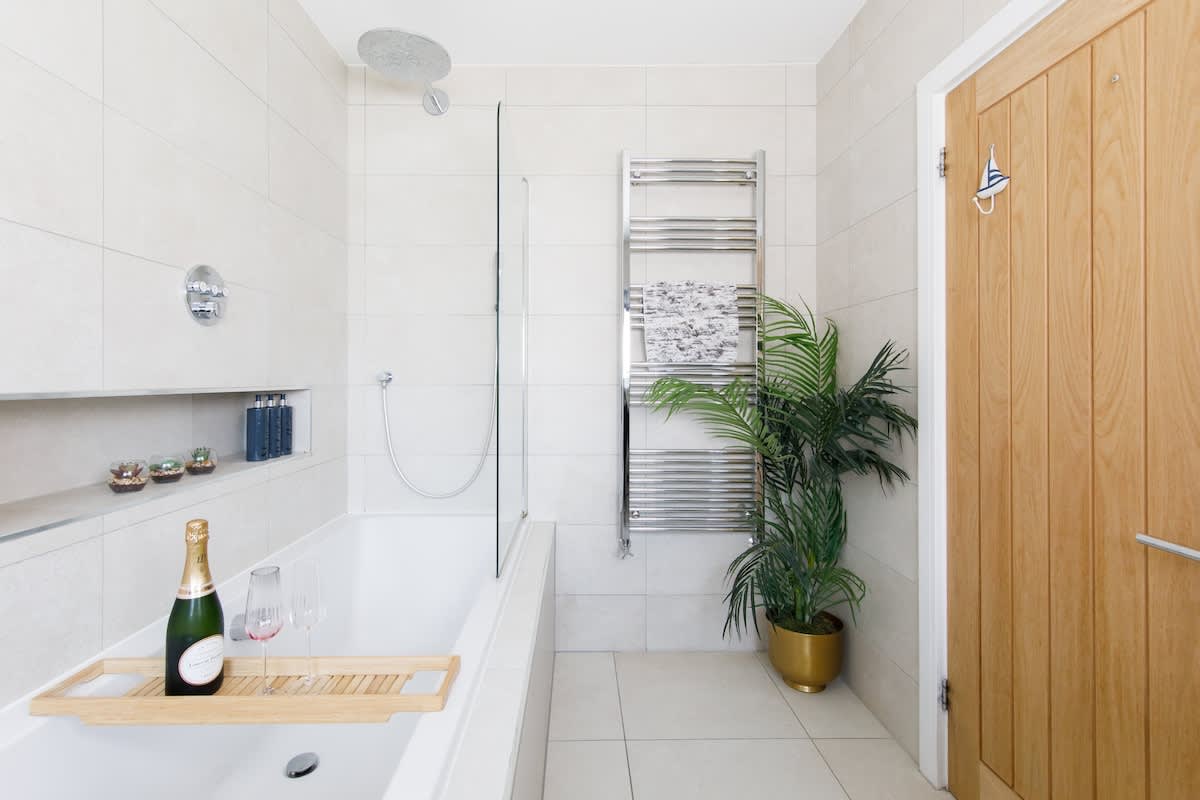 Cedar House: Elegant bathroom with a luxurious bathtub, rainfall shower, and heated towel rail. Features modern decor, greenery accents, and thoughtful touches like a bath tray, creating a serene space for relaxation