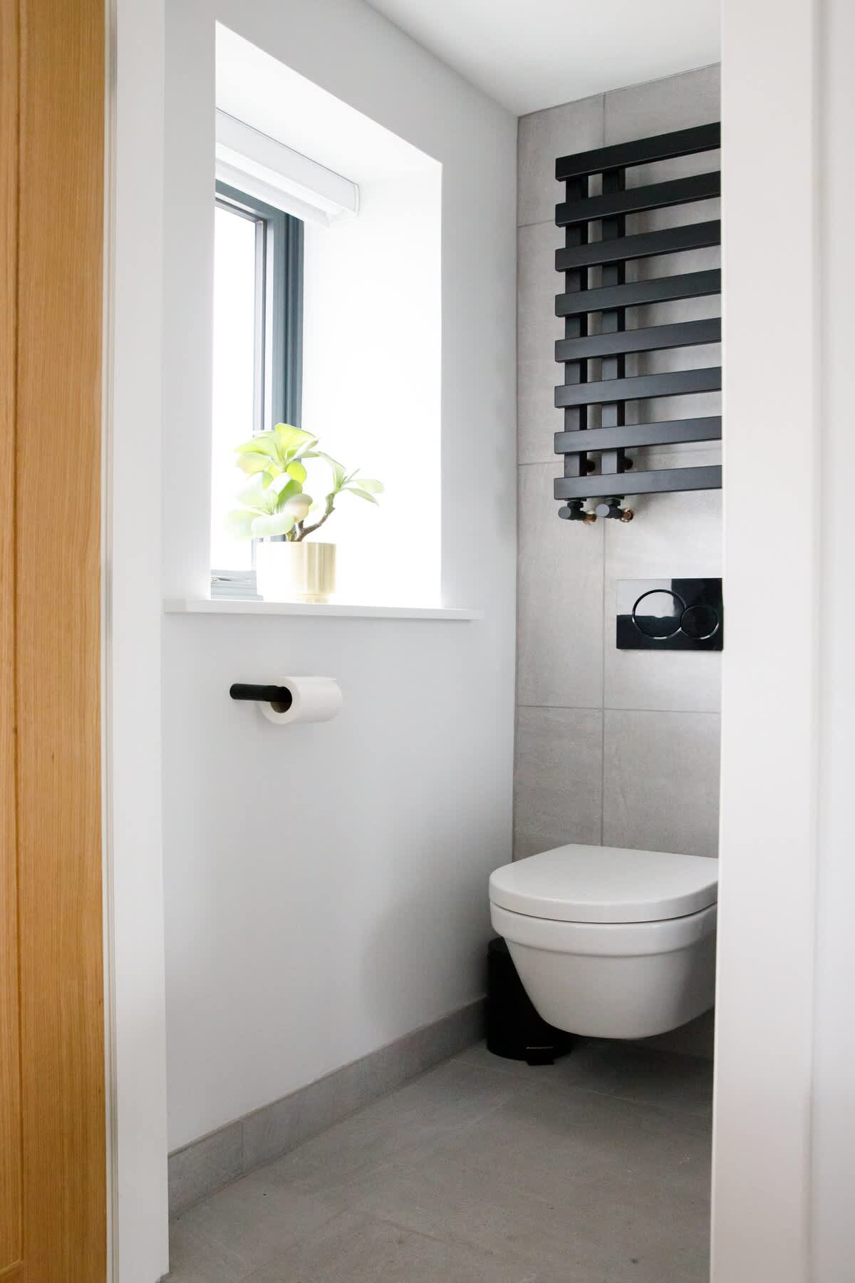 Cedar Lodge: Compact and modern WC featuring a wall-mounted toilet, sleek black heated towel rail, and natural light from a bright window. Minimalist design with thoughtful touches like a green plant for a clean and inviting space.