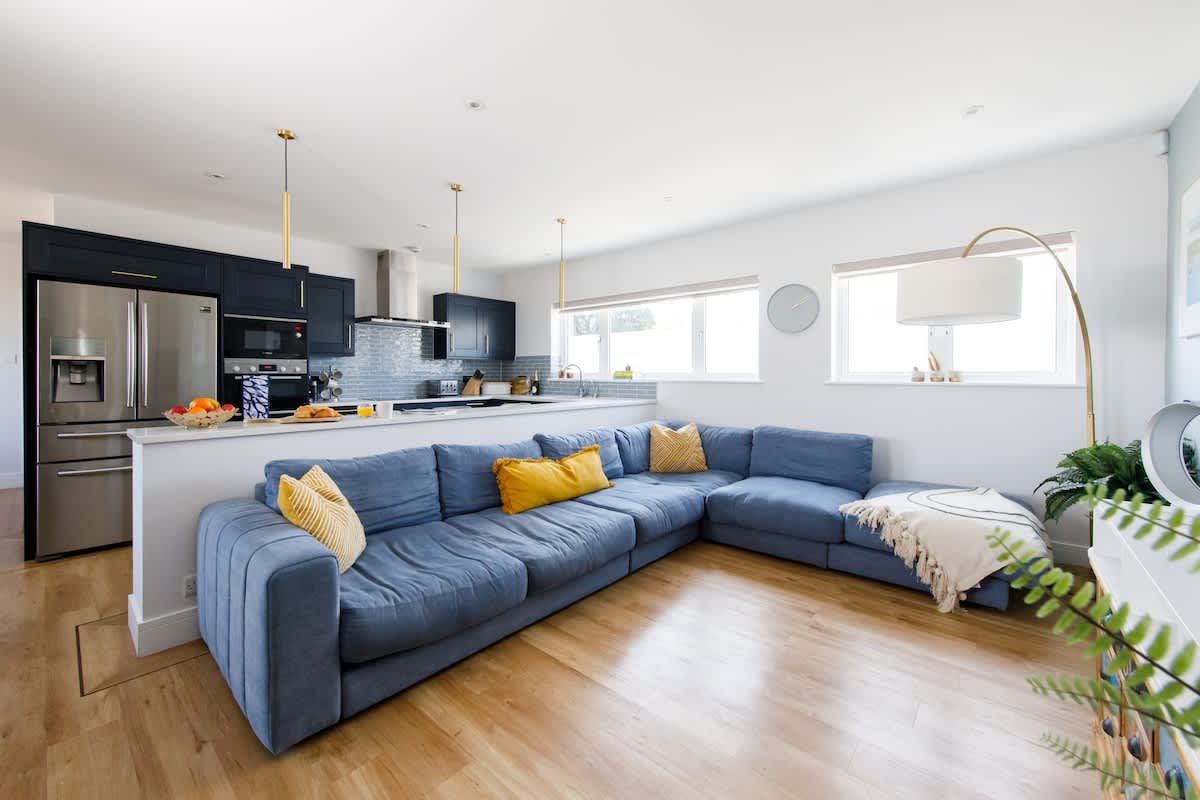 Bright & modern open-plan living space with a fully equipped kitchen, cozy blue sectional sofa, and stylish accents. Ideal for relaxing or entertaining, with natural light streaming in from large windows. Perfect for making your stay feel like home