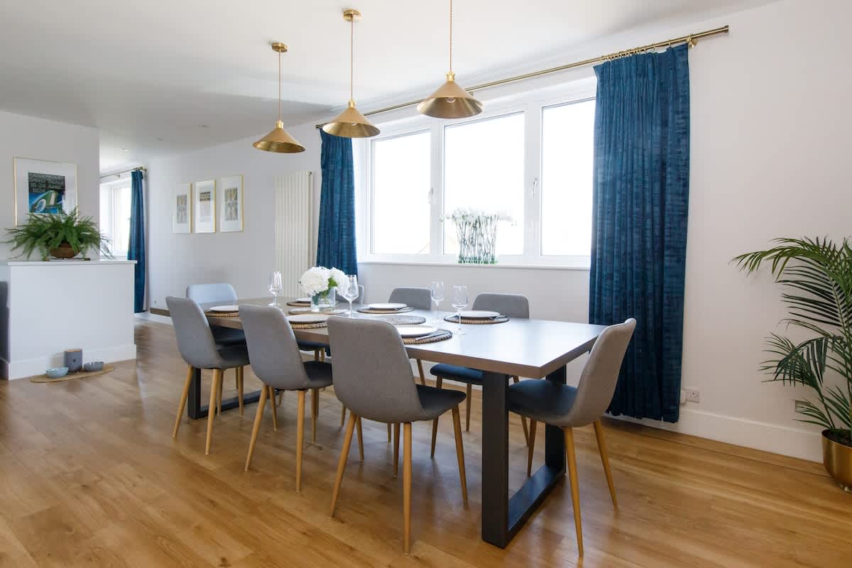 Elegant dining area with a sleek table seating 6, complemented by stylish chairs & gold-accented lighting. Natural light pours in through large windows framed by rich blue curtains, creating a cozy yet sophisticated atmosphere for meals & gatherings.