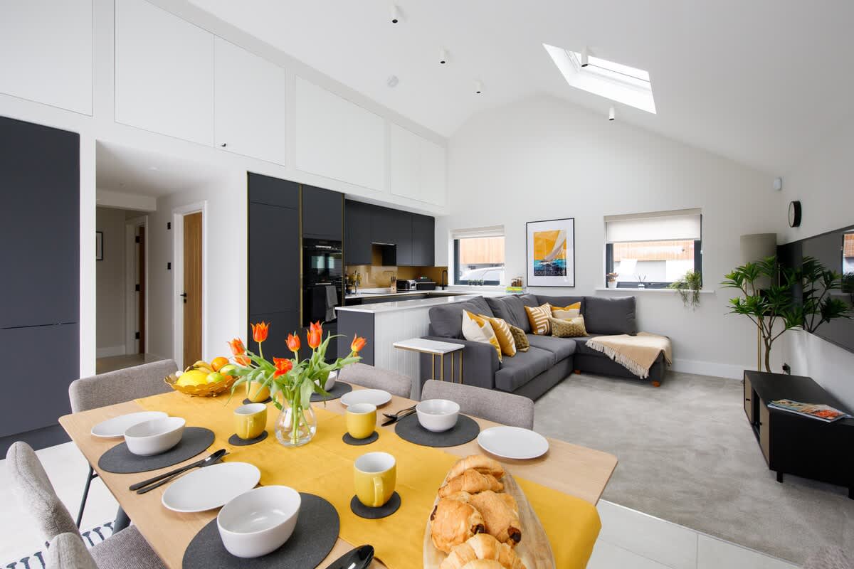 Cedar Lodge: Open-plan living and dining area with a stylish kitchen, cozy sectional sofa, and a beautifully set dining table. Skylights and large windows provide plenty of natural light, creating a bright and welcoming space for relaxation and meals