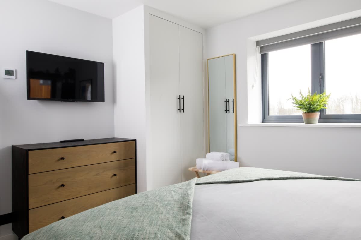 Cedar Lodge: Comfortable bedroom featuring a wall-mounted TV, spacious wardrobe, stylish dresser, and full-length mirror. Natural light from the window and tasteful decor create a bright and functional space for relaxation and convenience