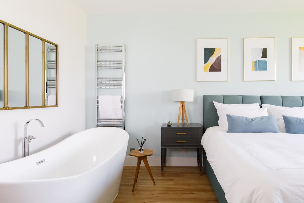 Bedroom 1 - Cedar House: Luxurious bedroom with a king-size bed, freestanding bathtub, and chic decor. Features modern artwork, a heated towel rail, and elegant furnishings, creating a serene retreat for ultimate relaxation and comfort.