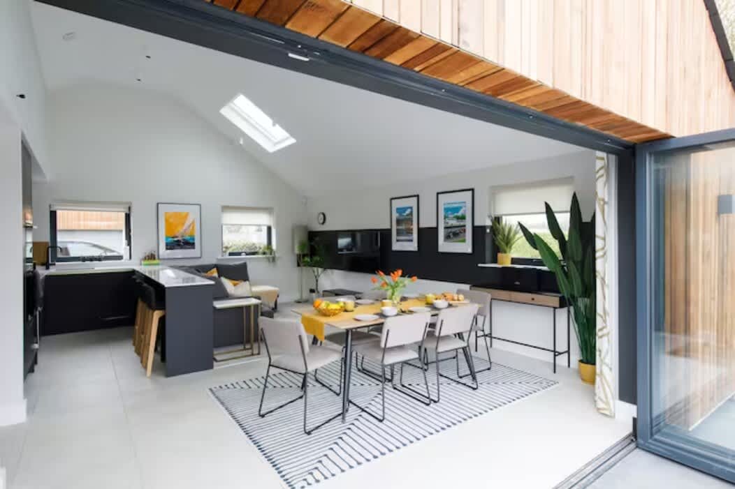 Bright open-plan kitchen & dining area with modern furnishings and bi-fold doors leading to the outdoors. Features a spacious dining table, skylight, & sleek design, perfect for meals or entertaining. Seamless indoor-outdoor flow for a relaxing stay