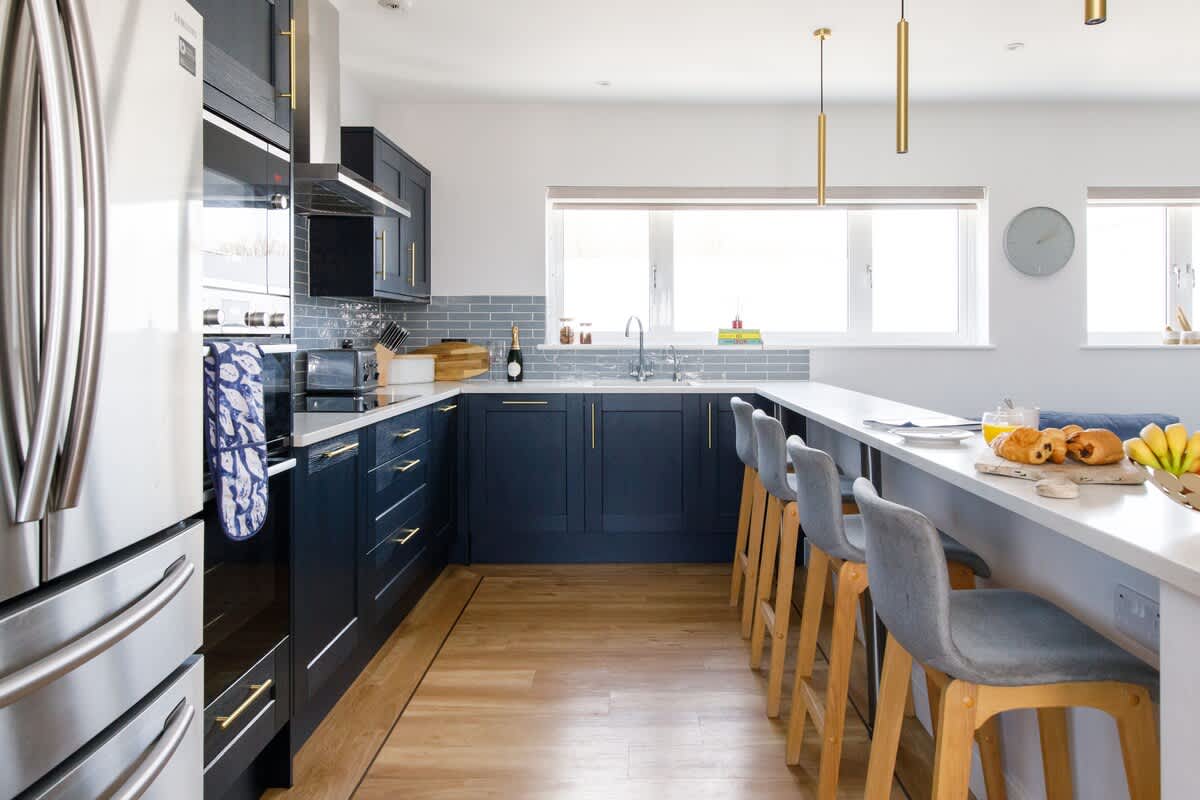 Cedar House: Stylish kitchen with navy cabinets, gold accents, and a breakfast bar with seating. Features stainless steel appliances, ample counter space, and natural light, making it a perfect space for cooking and socializing.
