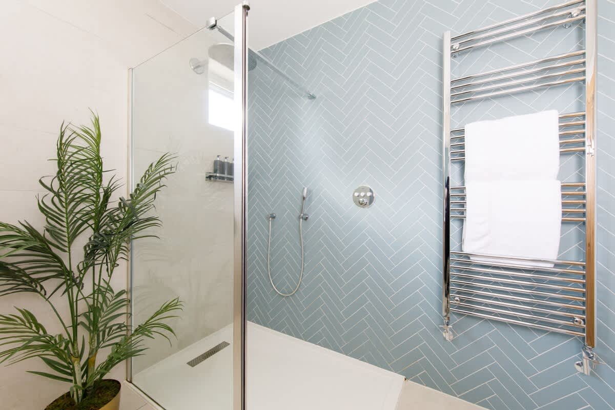 Cedar House: Modern walk-in shower with stylish blue herringbone tiles, a glass enclosure, and a sleek rainfall showerhead. Features a heated towel rail and greenery accents, creating a fresh and luxurious bathroom experience