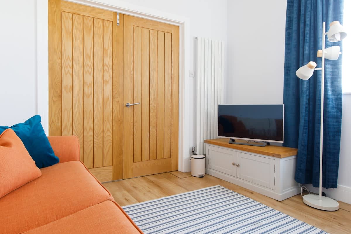 Bedroom 4 - Cedar House: Bright and inviting lounge area with a cozy orange sofa, flat-screen TV, and striped rug. Perfect for unwinding or enjoying family time, with natural wood accents and a stylish floor lamp completing the space.