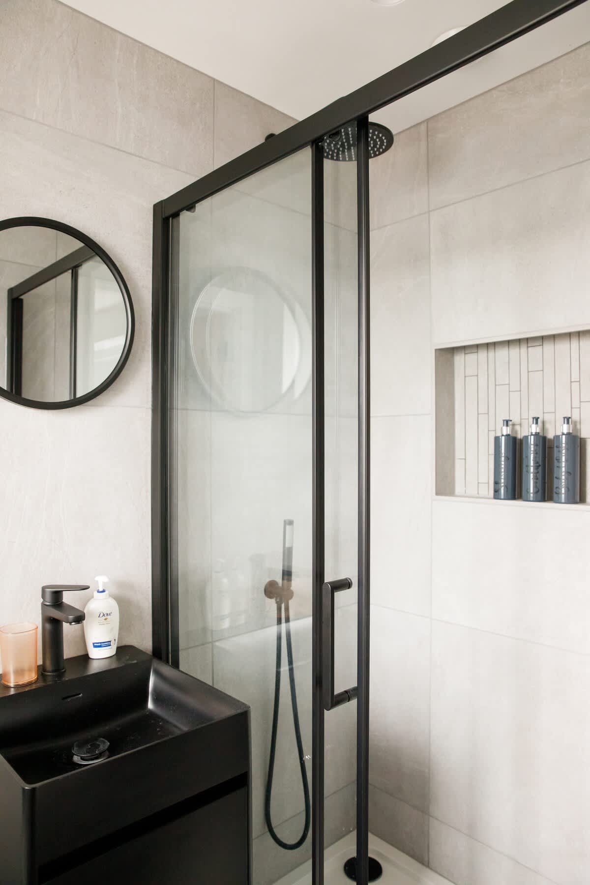 Cedar Lodge: Modern bathroom with a sleek black vanity, glass-enclosed shower, and rainfall showerhead. Includes built-in shelving with amenities, minimalist decor, and stylish finishes, offering a luxurious and functional space for your stay.