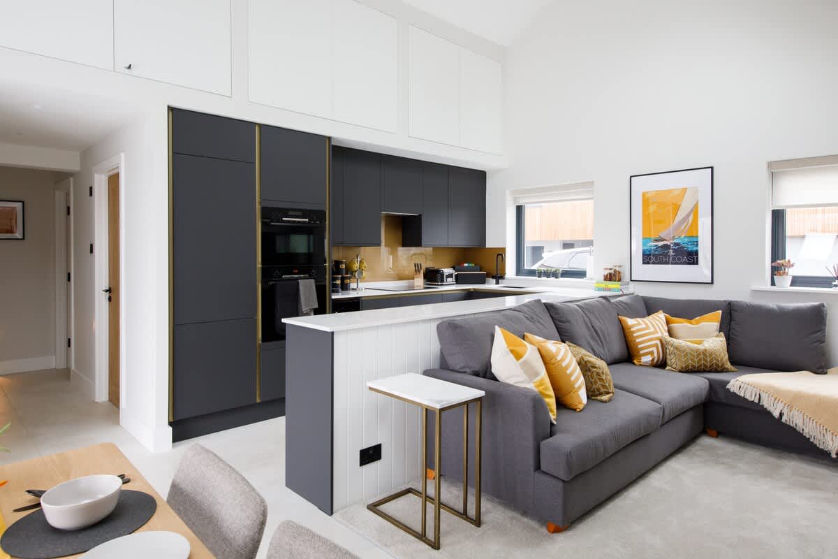 Cedar Lodge: Modern open-plan living area featuring a sleek kitchen with high-end appliances and a cozy sectional sofa with vibrant accents. Perfect for relaxing or entertaining, with natural light streaming in through large windows.