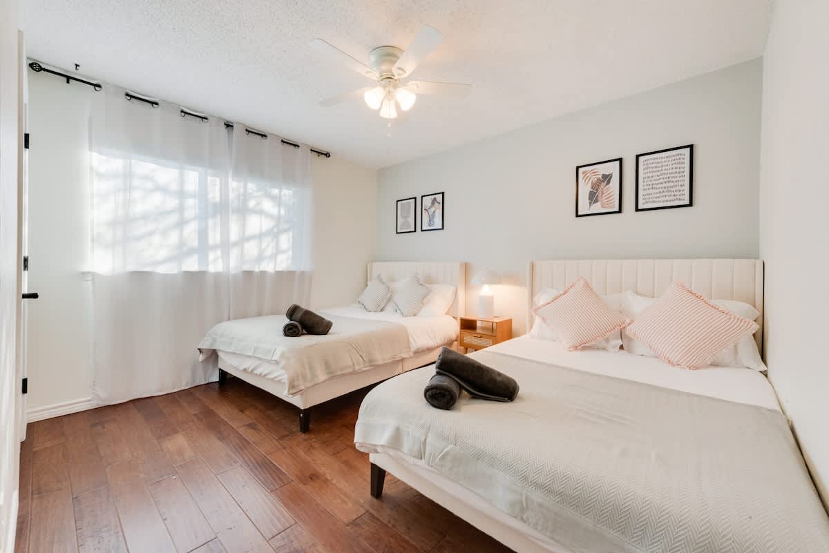 This bright and cozy bedroom features 2 full beds, accommodating up to 4 guests. It boasts soft decor w/ neutral tones, and a ceiling fan for comfort. A large window with sheer curtains lets in natural light, creating a warm and inviting atmosphere.
