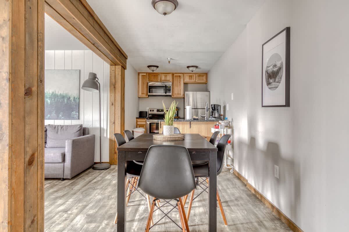 Dining area with a dark rectangular table and six black modern chairs, leading into a compact kitchen with light wood cabinetry, stainless steel appliances, and a breakfast bar.