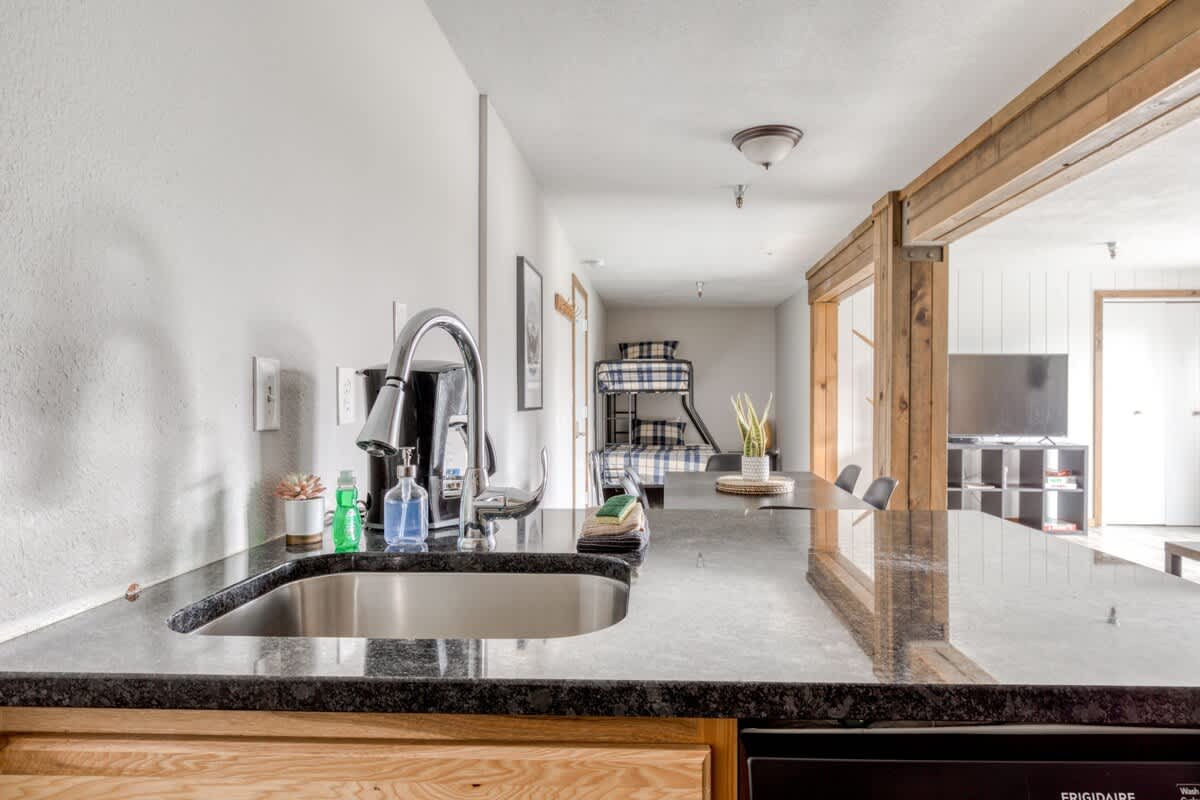 A kitchen island with a dark granite countertop and a stainless steel sink, looking into an open-concept space with a dining table and a bunk bed visible in the background, featuring light wood accents.