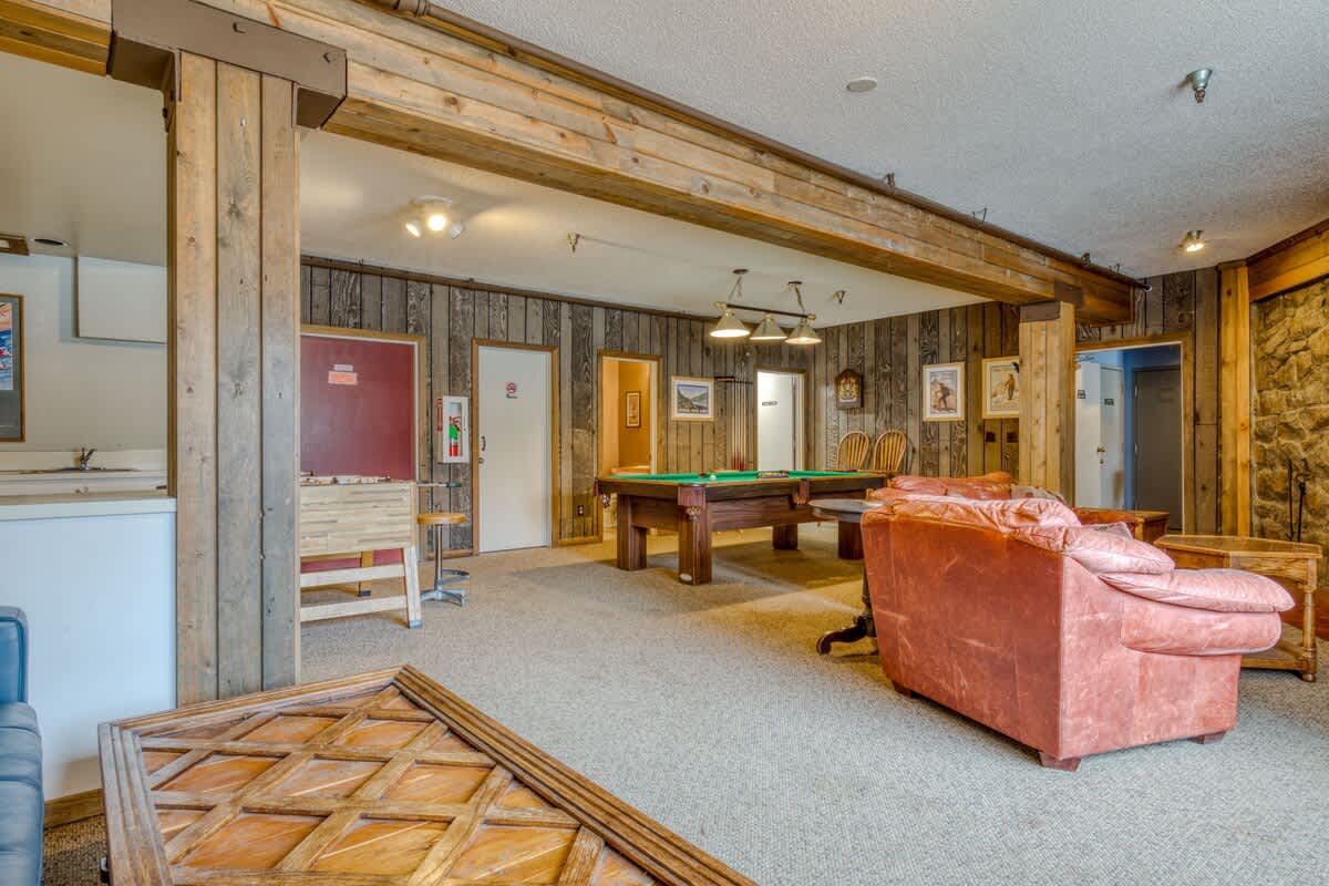 A spacious communal game room with wood-paneled walls, exposed wooden beams, and carpeted flooring, featuring a large pool table, a foosball table, and comfortable seating. The room has a rustic, lodge-like aesthetic.