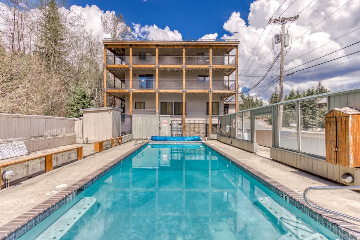 Outdoor rectangular swimming pool with bright blue water, surrounded by concrete decking and a multi-story building with wooden balconies. Lush trees and a utility pole are visible in the background under a partly cloudy sky.