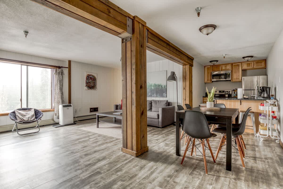 Open-concept living space with rustic wooden beams separating the living room, dining area, and kitchen, featuring light laminate flooring and a mix of modern and cozy furnishings.