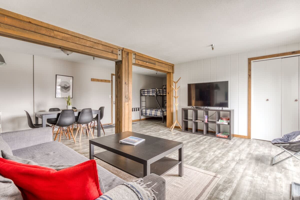 Spacious living room featuring a gray sofa with a red accent pillow, a dark coffee table, and a view towards the dining area and kitchen, with a bunk bed nook to the right.
