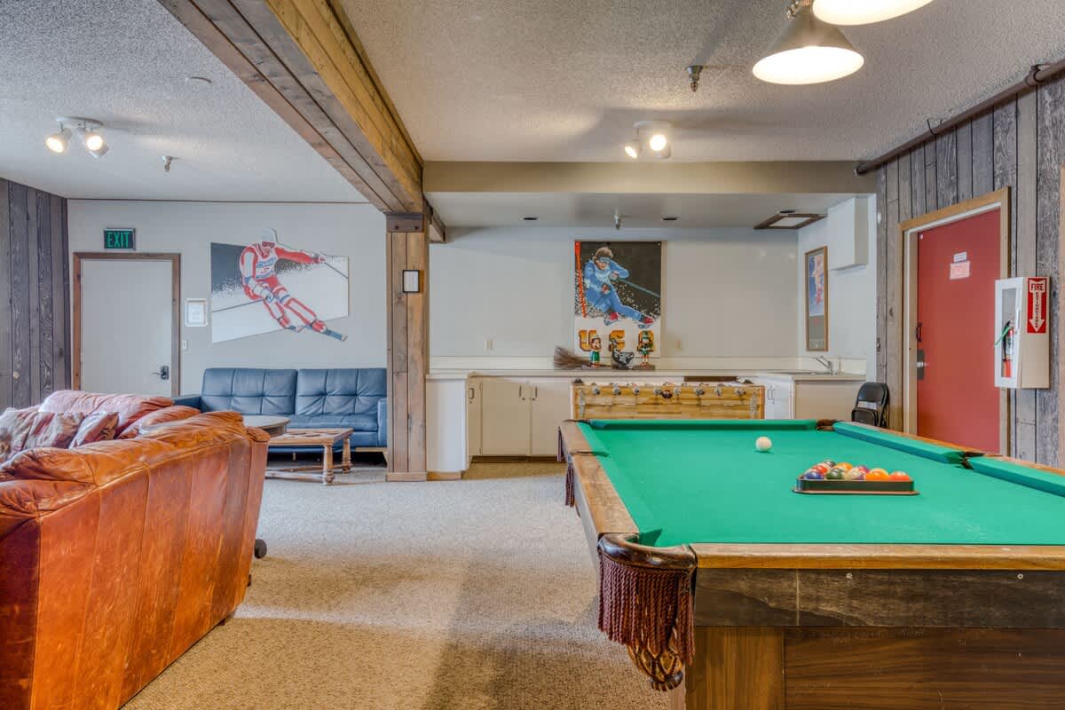 Spacious game room featuring a green felt pool table as the centerpiece, alongside a foosball table and various seating options including a blue sofa and a brown leather couch. Ski-themed artwork adorns the paneled walls, creating a recreational atmosphere.