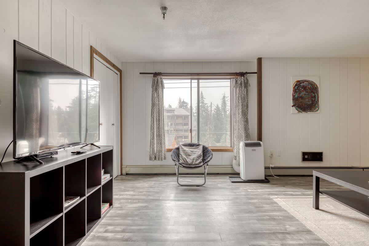 Bright living room featuring white vertical paneling, a large flat-screen TV on a dark shelving unit, and a cozy armchair by a window with patterned curtains and a portable AC unit.