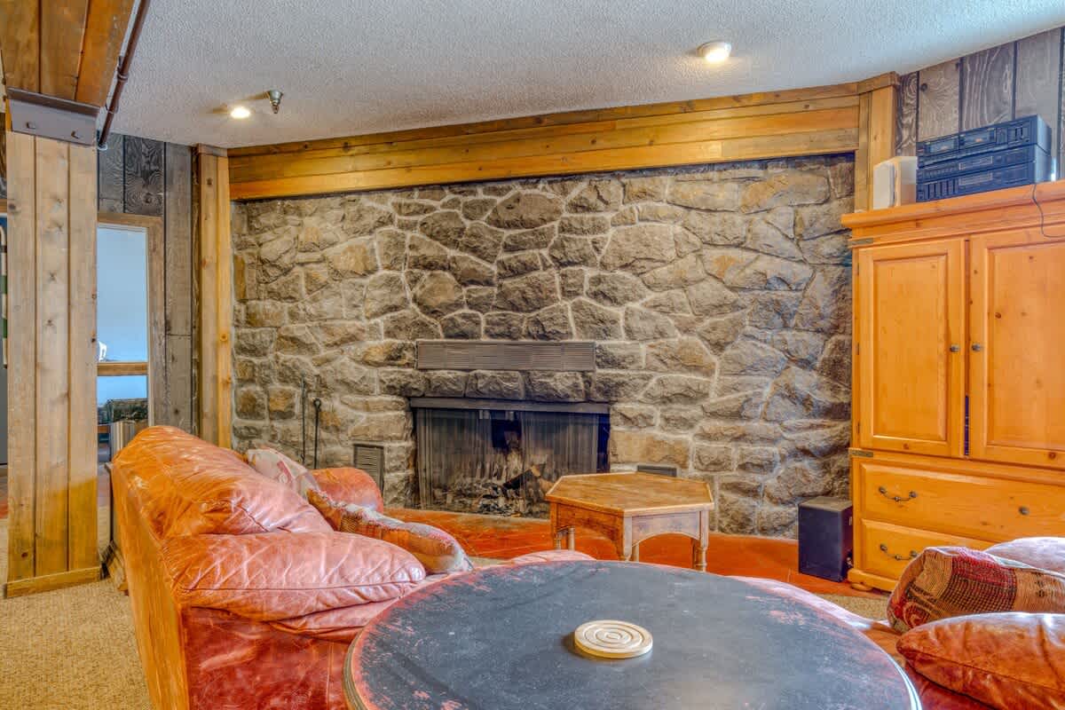 A rustic living room centered around a large stone fireplace with a wooden mantel, furnished with a comfortable leather sofa and a round wooden coffee table, all set on a carpeted floor.