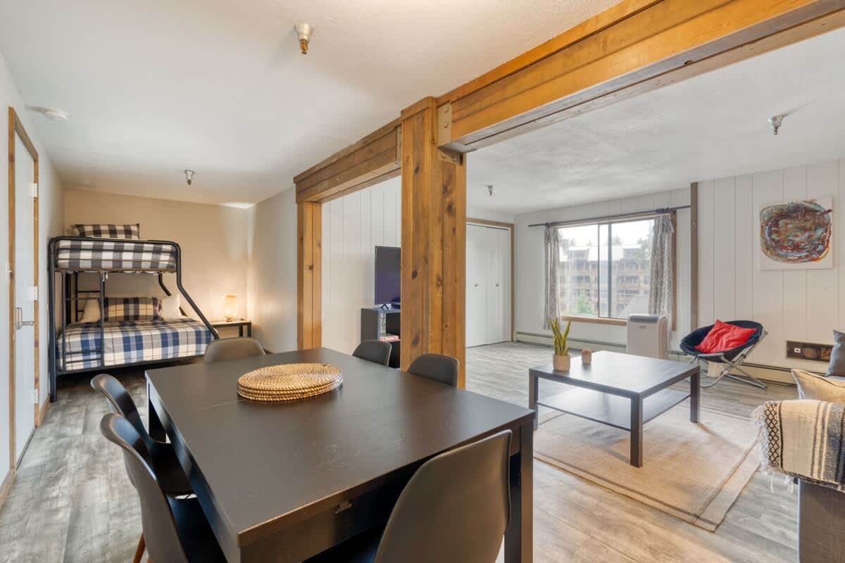 Expansive view of the open-concept living space, featuring a dark dining table, a comfortable living area with a sofa, and a distinct bunk bed sleeping nook, all connected by rustic wooden beams.