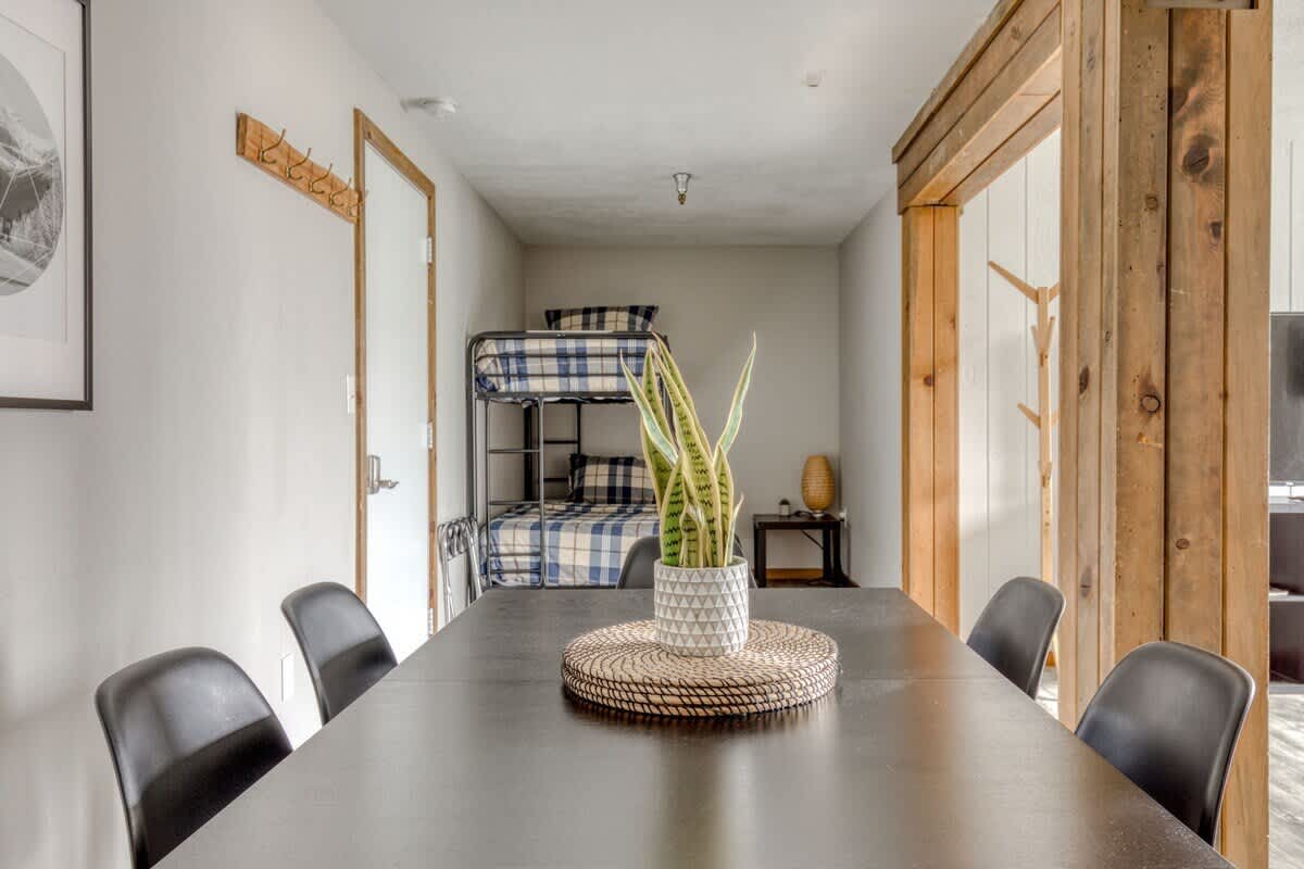A view from the dining area towards a hallway, revealing a bunk bed sleeping area in the distance, with a dark dining table and chairs in the foreground and a potted snake plant as a centerpiece.