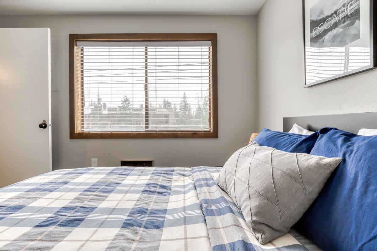 A close-up of a comfortable queen bed with blue and white plaid bedding, accented with blue and gray throw pillows, set against a dark headboard and a light gray wall.