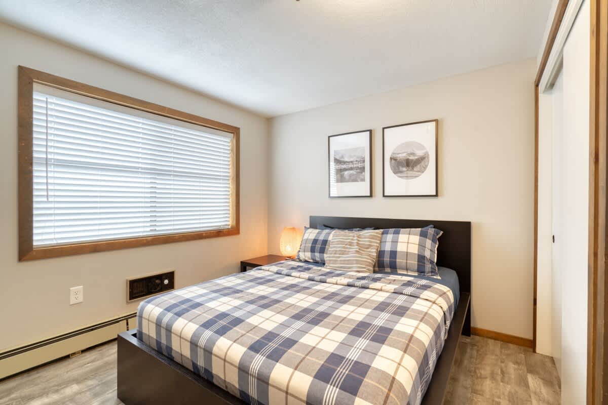 A bright bedroom featuring a queen bed with blue and white plaid bedding and a dark frame, positioned beneath a large window with wooden blinds and a wooden frame, complemented by two framed art pieces on the wall.