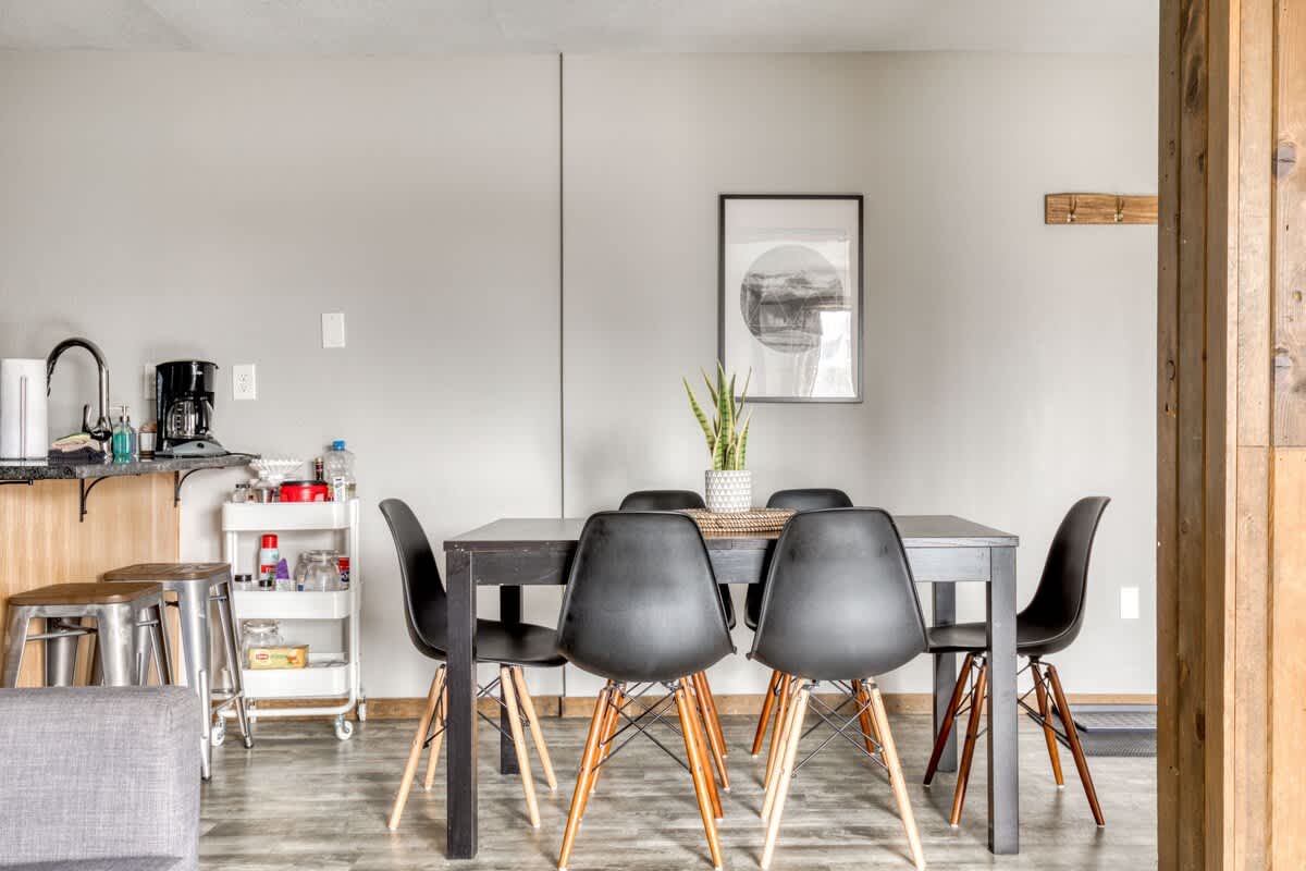 A modern dining area with a dark rectangular table surrounded by six black chairs with light wood legs, centered under a framed abstract art piece on a light gray wall, with a snake plant as a centerpiece.