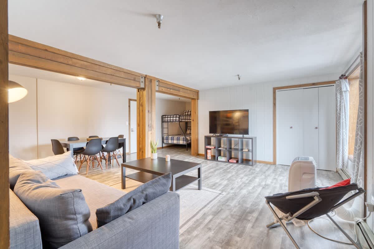 Wide view of the living room, showing the gray sofa, coffee table, and TV area. The bunk bed alcove is clearly visible to the left, and the open layout connects to the dining area through rustic wooden beams, all on light wood-look flooring.