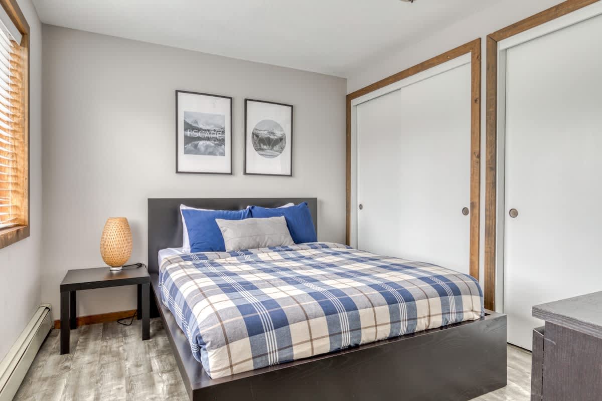 Cozy bedroom with a queen-sized bed featuring a blue and white plaid comforter, flanked by a dark nightstand with a textured lamp. Two framed prints hang above the bed, and white sliding closet doors with wooden trim are visible.