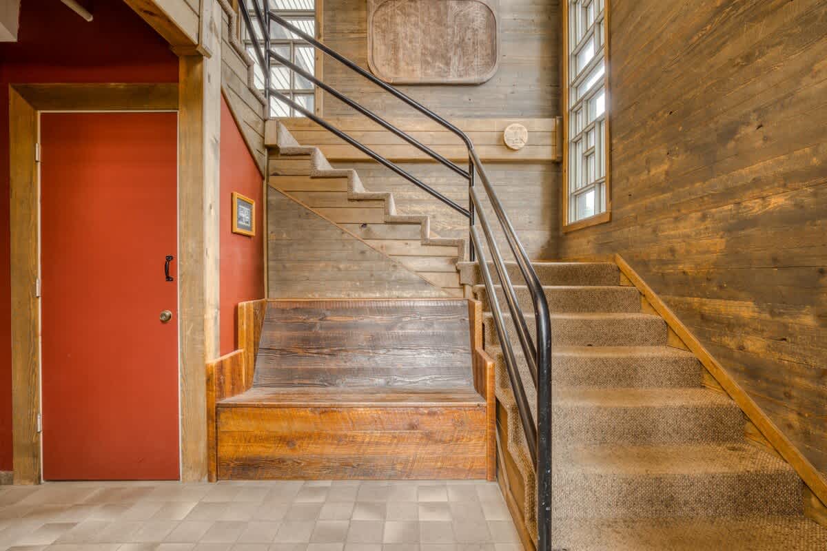 A rustic interior space featuring a wooden bench, a red door, and a carpeted staircase with a black metal railing, all set against wood-paneled walls with tall, multi-pane windows, creating a cozy lodge ambiance.