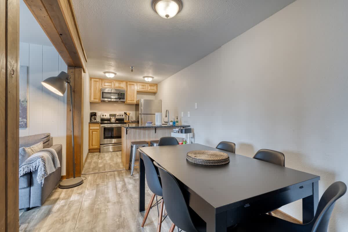 Dining area with a dark rectangular table and black chairs, adjacent to a compact kitchen featuring light wood cabinets, stainless steel appliances, and a small island with bar stools.