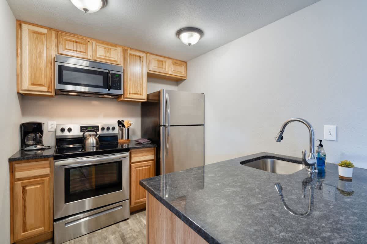 A well-equipped kitchen featuring light wood cabinetry, dark granite countertops, and stainless steel appliances including a microwave, range, and refrigerator, with a coffee maker and kettle on the counter.