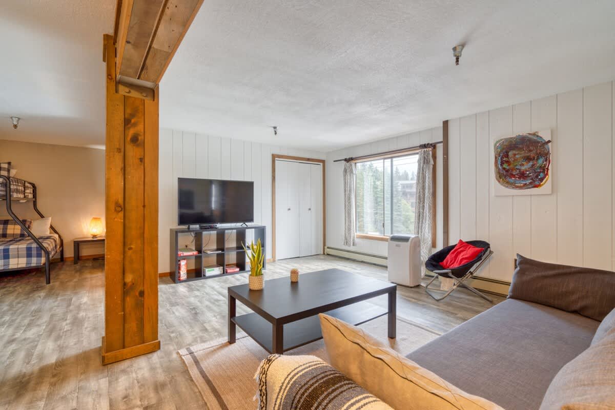 Living room featuring a gray sofa, dark coffee table, and a large wall-mounted TV on a black media console. A bunk bed is visible in an alcove to the left, separated by wooden beams, all on light wood-look flooring.