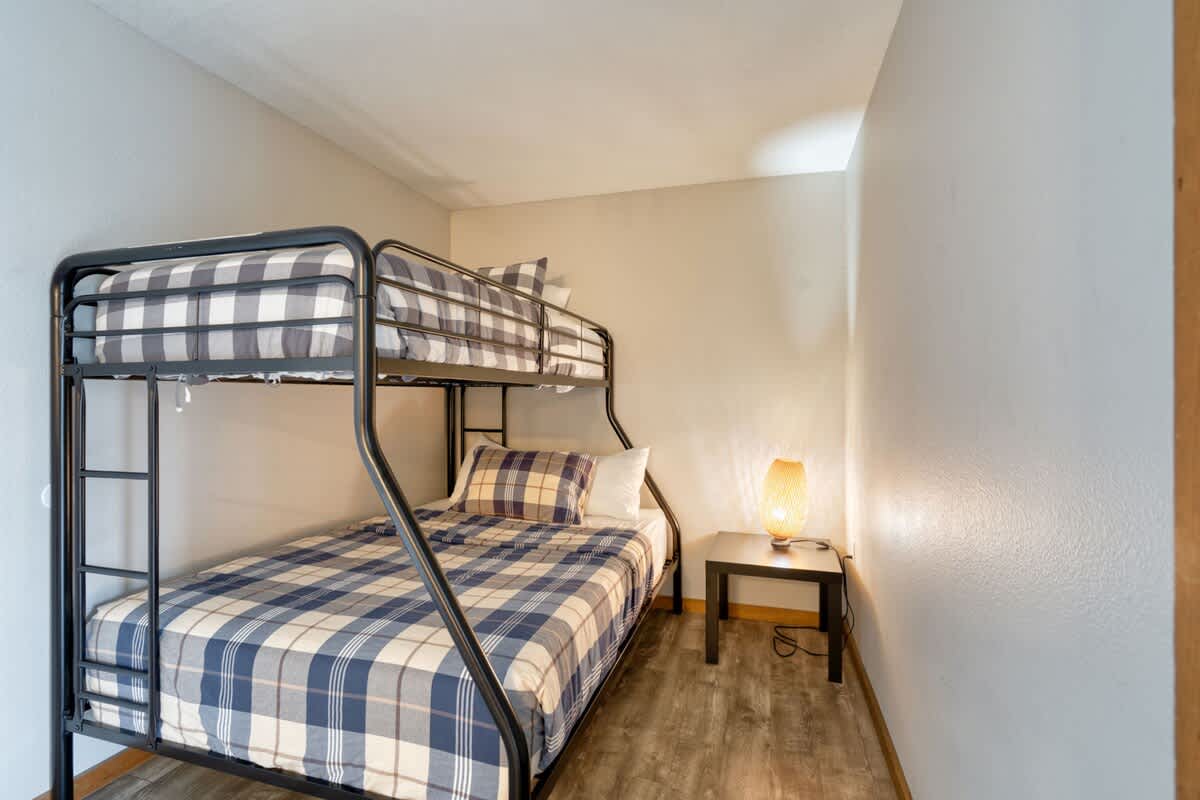 A compact sleeping nook featuring a black metal bunk bed with blue and white plaid bedding, a small wooden nightstand, and a lamp with an orange textured shade, all set against light gray walls and wood-look flooring.