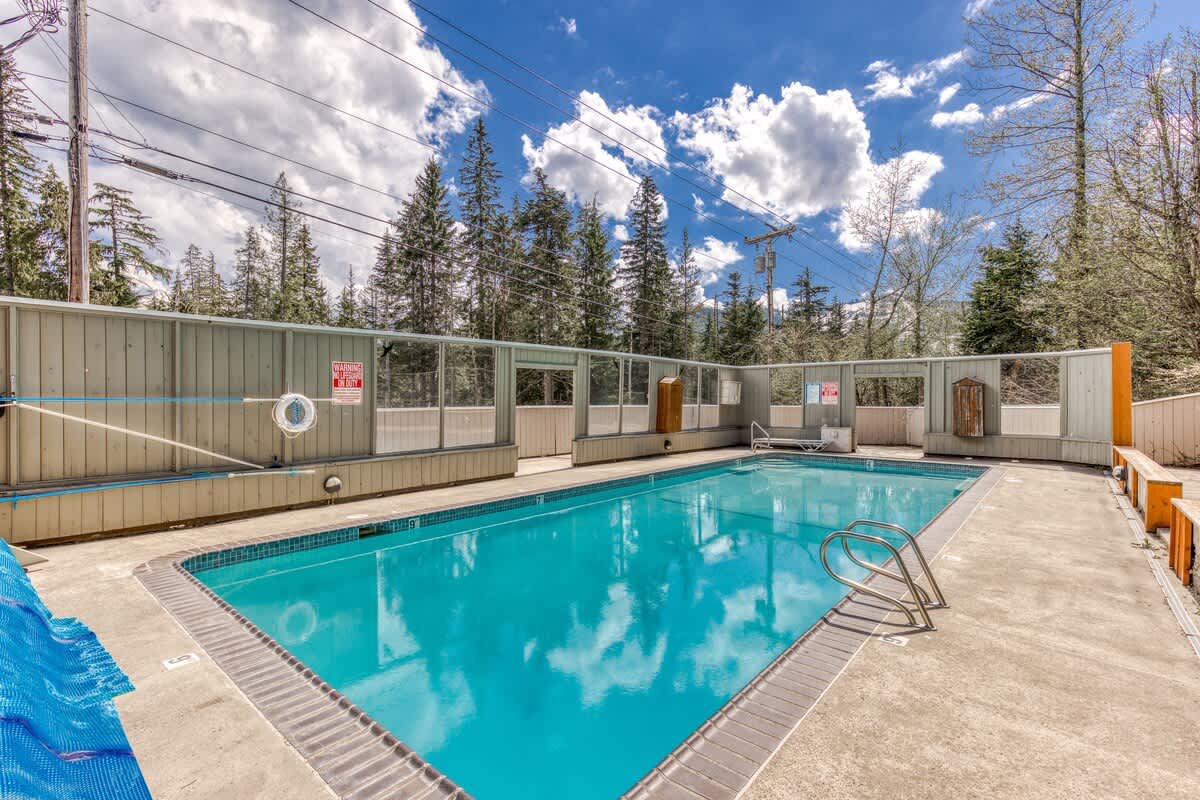 An outdoor swimming pool with clear blue water, surrounded by a concrete deck and a low fence, with tall trees and power lines visible in the background under a partly cloudy sky. A blue pool cover is folded on the left.