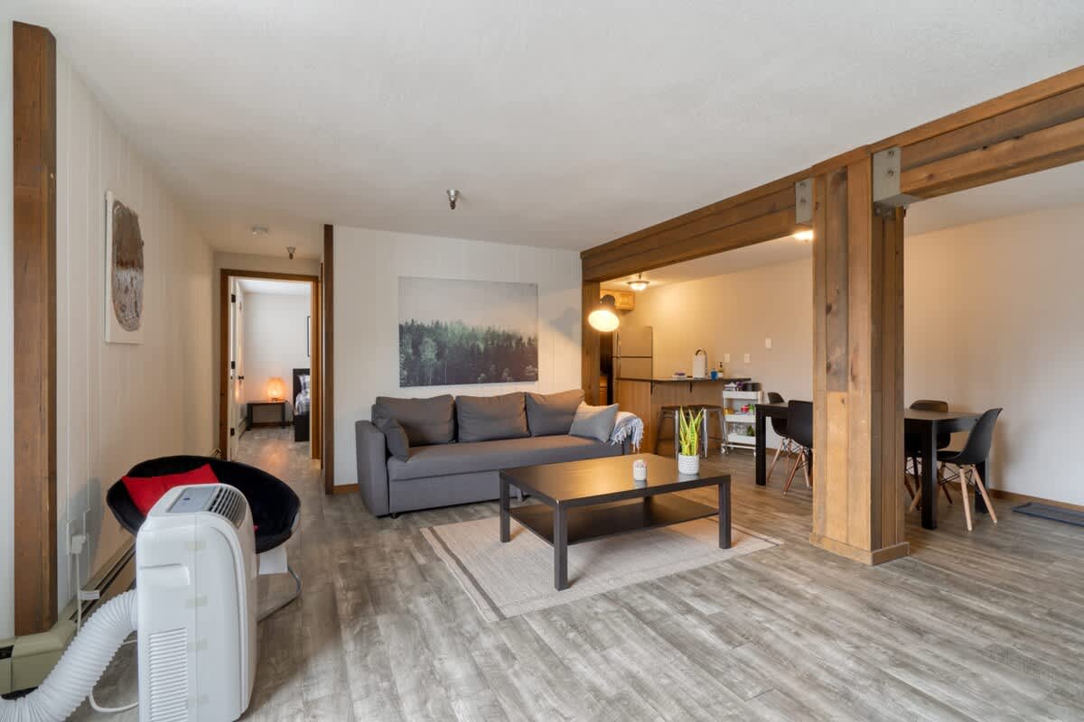 Living room with a gray sofa, dark coffee table, and a portable air conditioner by the window. A doorway leads to another room, and the light wood-look flooring extends throughout the bright, open space with white walls.