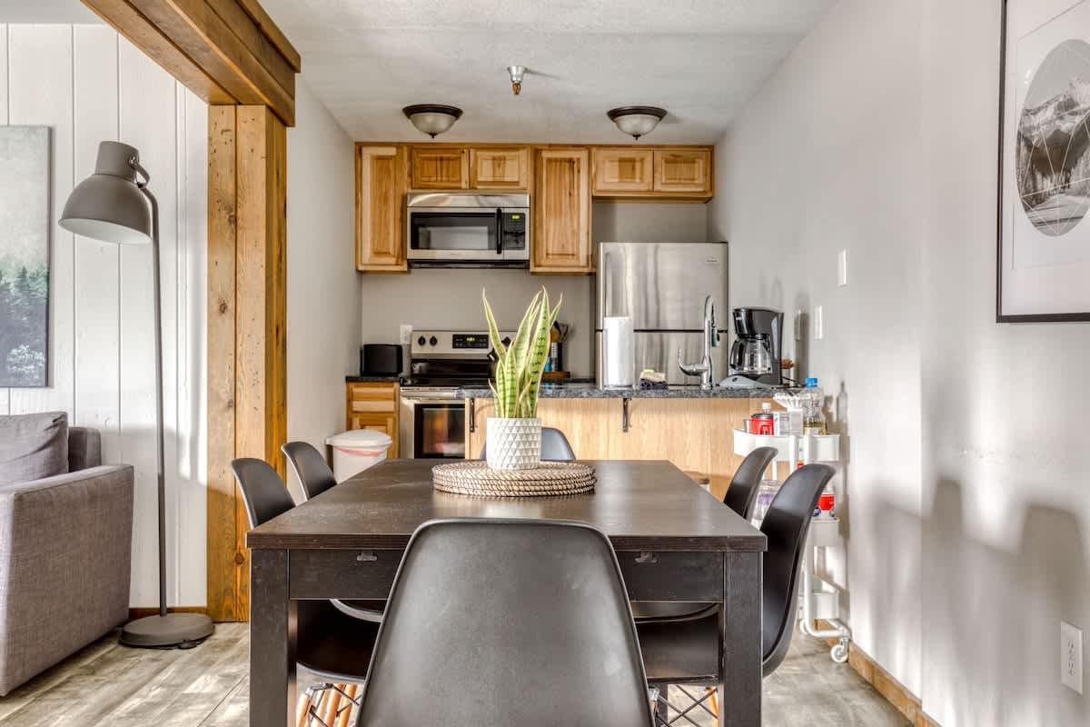 Open-concept dining area featuring a dark wooden table with black chairs, adjacent to a compact kitchen. The kitchen has light wood cabinets, stainless steel appliances, and a breakfast bar with stools, all framed by rustic wooden beams.