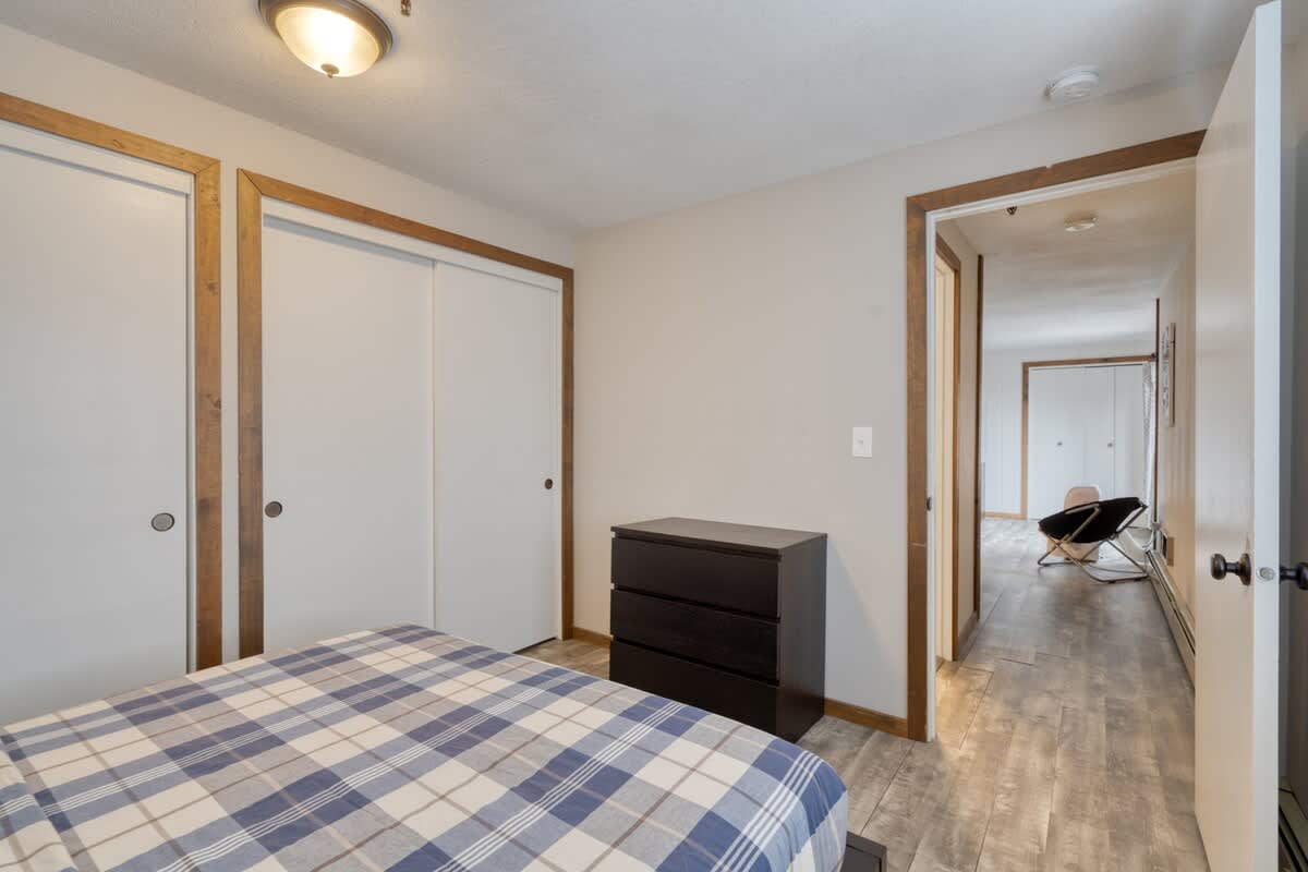 A view inside bedroom-1 showing a dark wooden dresser and two white sliding closet doors with wooden frames, looking out into a hallway with wood-look flooring and another room visible in the distance.