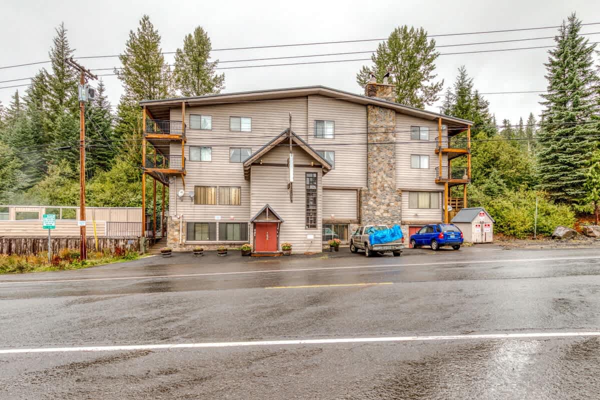 A multi-story lodge-style building with light brown siding and a prominent stone chimney, surrounded by tall evergreen trees under an overcast sky. Two cars are parked in front, and balconies are visible on the upper floors.