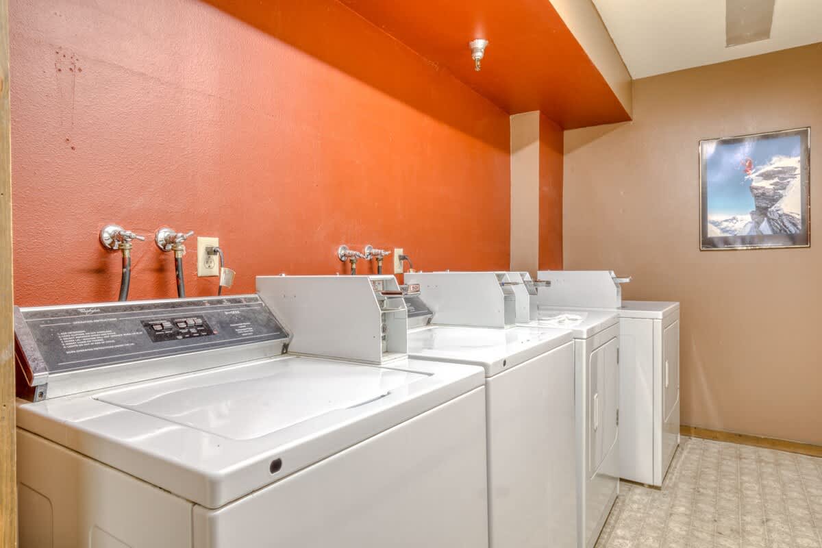 A functional laundry room with several white top-loading washing machines and dryers lined against a vibrant orange and beige wall. The room has a tiled floor and a framed picture on the wall.