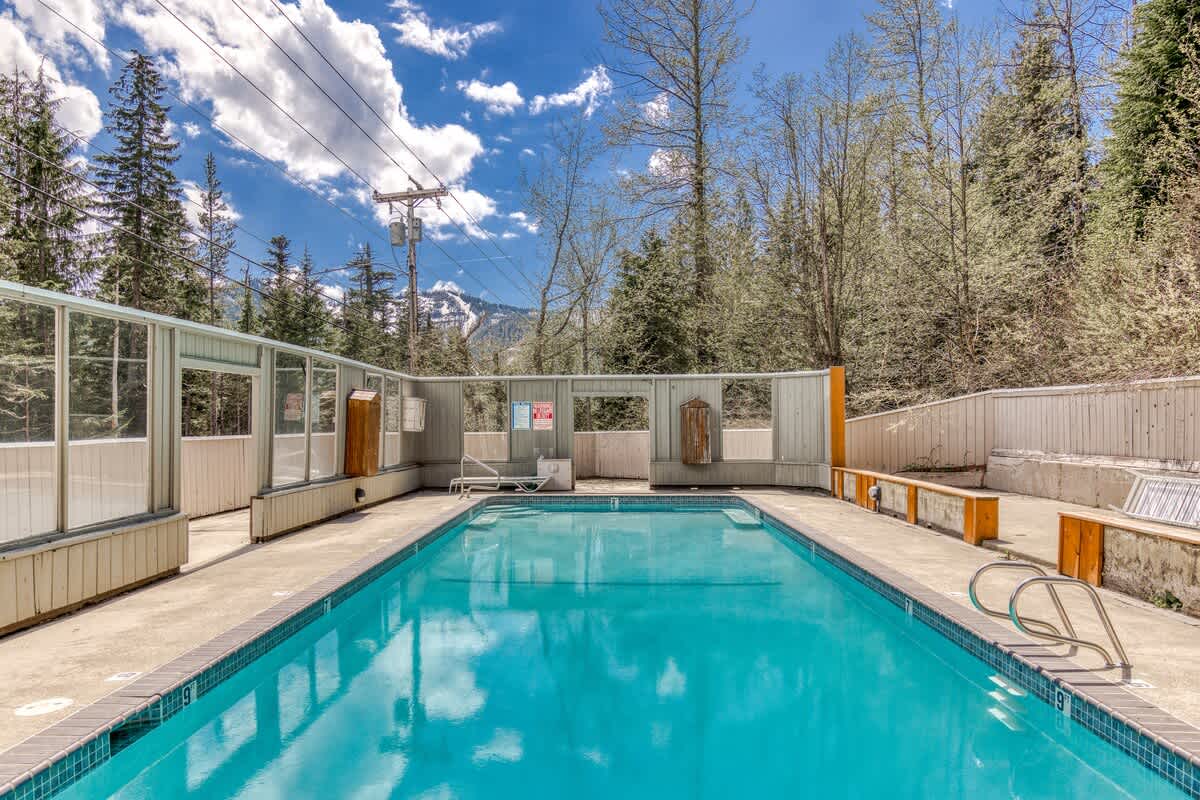 A wide shot of the outdoor swimming pool, showing the surrounding concrete patio, a low fence with glass panels, and benches, all set against a backdrop of lush green trees and a bright blue sky with white clouds.