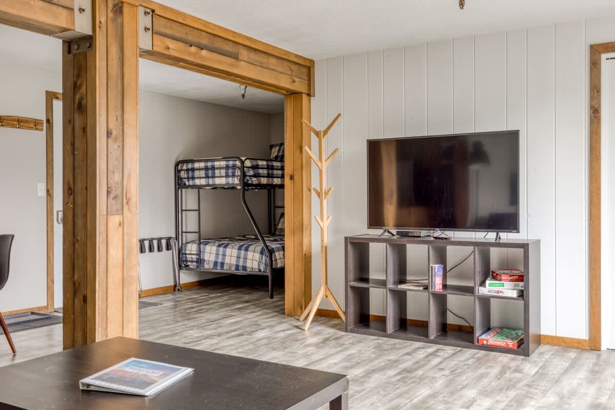 Living room with a large flat-screen TV on a dark shelving unit, a dark coffee table, and a wooden coat rack, with a view into a sleeping nook featuring bunk beds.