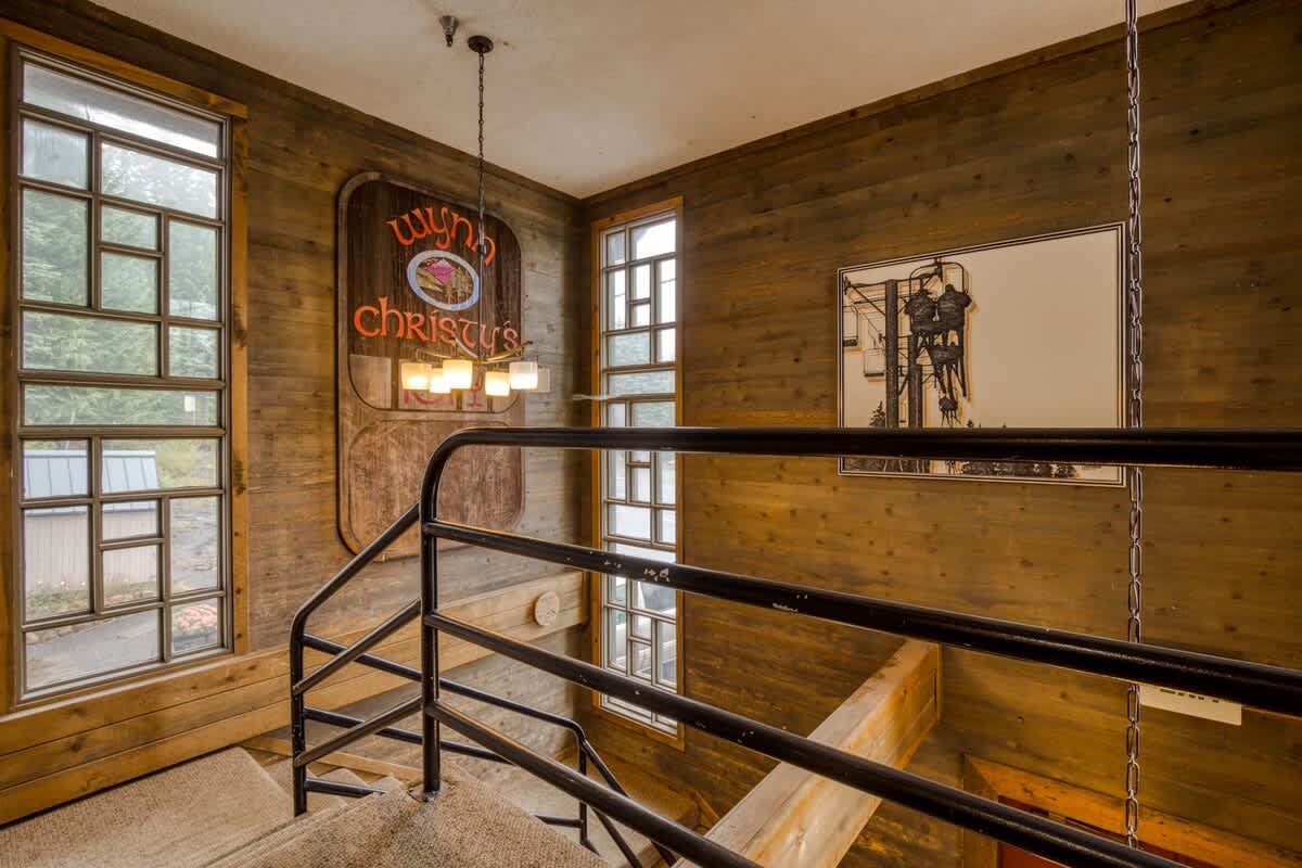 A view from the top of a carpeted staircase with a black metal railing, showcasing wood-paneled walls adorned with a large vintage "Wynn's Christies" sign and a framed ski lift illustration, illuminated by a hanging chandelier and tall windows.
