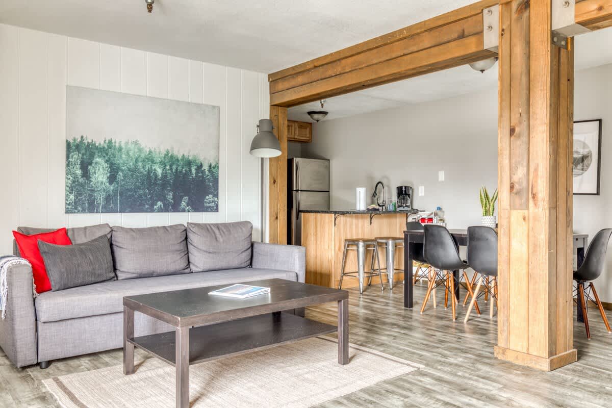 Wide shot of the open-concept living area, featuring a gray sofa and coffee table in the foreground. The kitchen and dining area are visible beyond the rustic wooden support beams, all unified by light wood-look flooring.